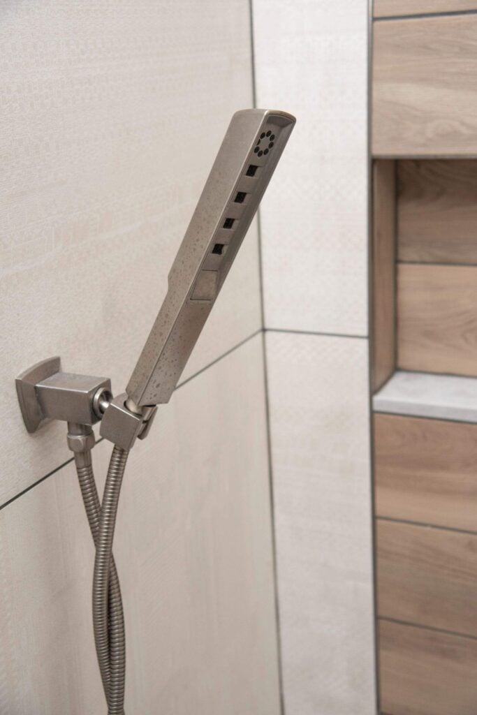 A modern handheld showerhead with a rectangular design is mounted on a tiled bathroom wall, with a flexible metal hose attached. Wooden shelves are visible in the background.