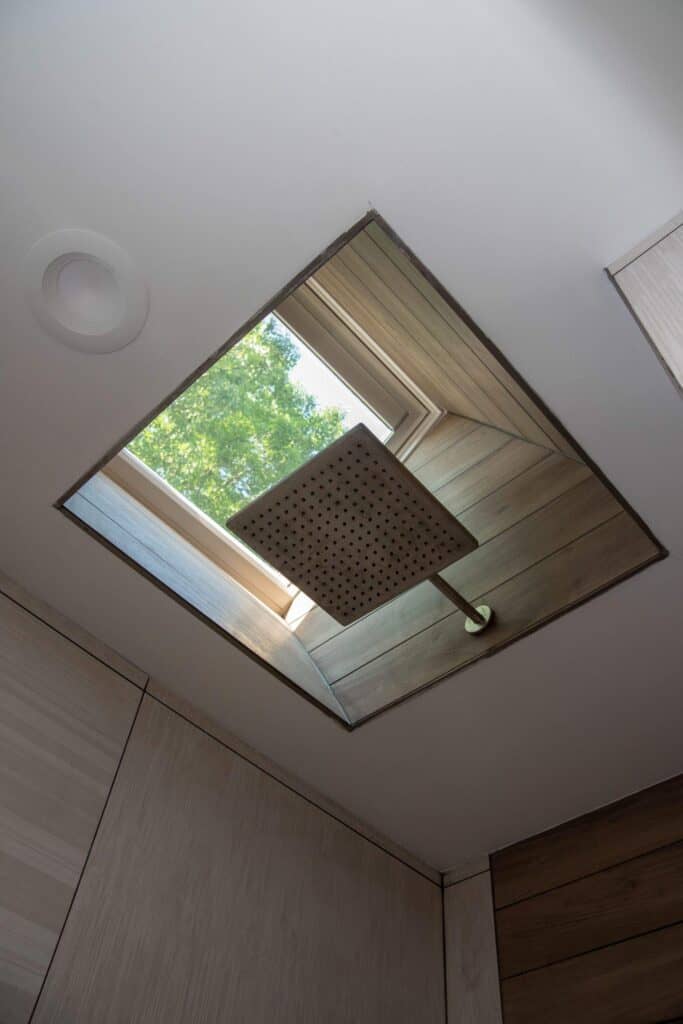 A square rainfall showerhead is mounted directly below a skylight window on a wooden ceiling, with green tree foliage visible through the open skylight. The shower is surrounded by light-colored tiled walls.