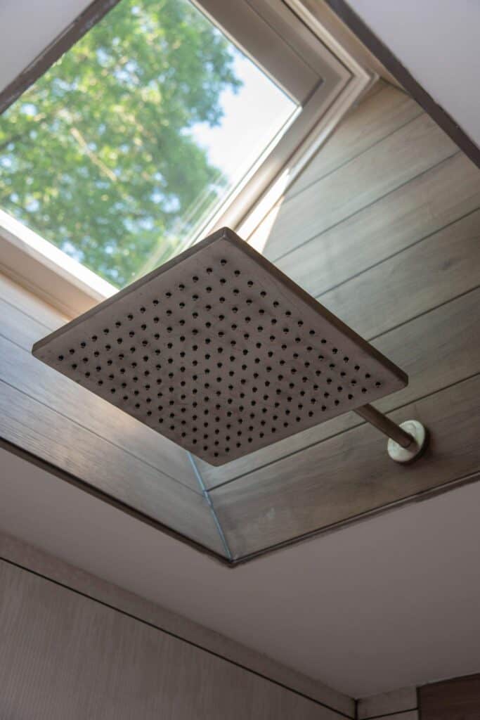 A square showerhead is mounted on the ceiling beneath a large skylight window, allowing natural light to enter the bathroom. Trees and blue sky are visible through the window.