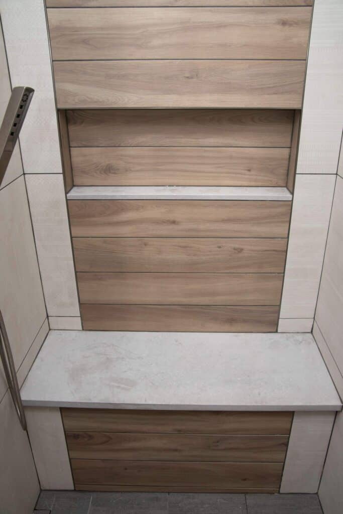 A shower area with beige and wood-look tile walls, featuring a built-in bench with a light stone top and two recessed shelves above, all framed by neutral-toned tiles.