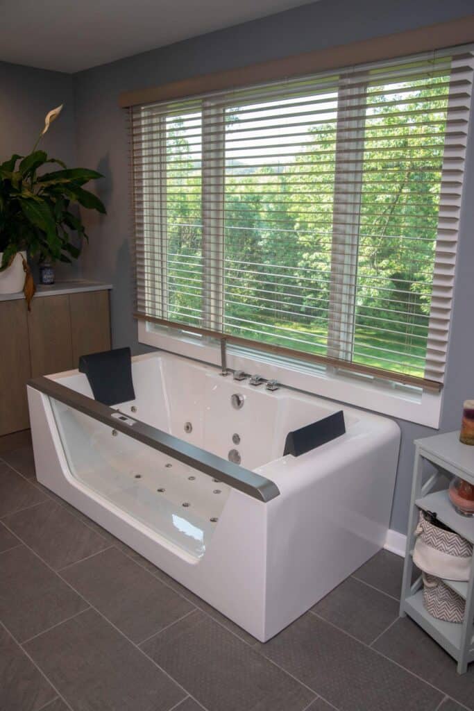 A modern white bathtub with black headrests and built-in jets sits beneath large windows with blinds, overlooking a green outdoor view. The bathroom has gray tile floors and light blue walls.