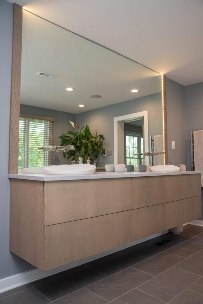 A modern bathroom with a large mirror above a floating wood vanity, two vessel sinks, and minimalist fixtures. A potted plant sits between the sinks, and natural light streams in from a window.