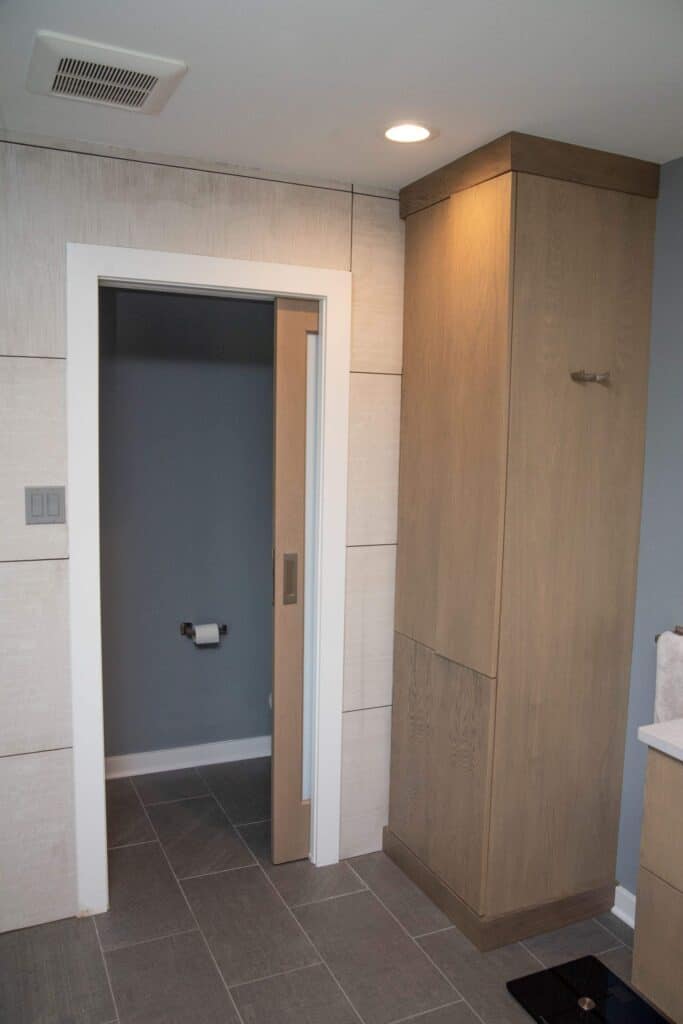 A modern bathroom with gray tile flooring, a tall wooden cabinet, and a sliding door leading to a small room with a toilet and a toilet paper holder on the wall.