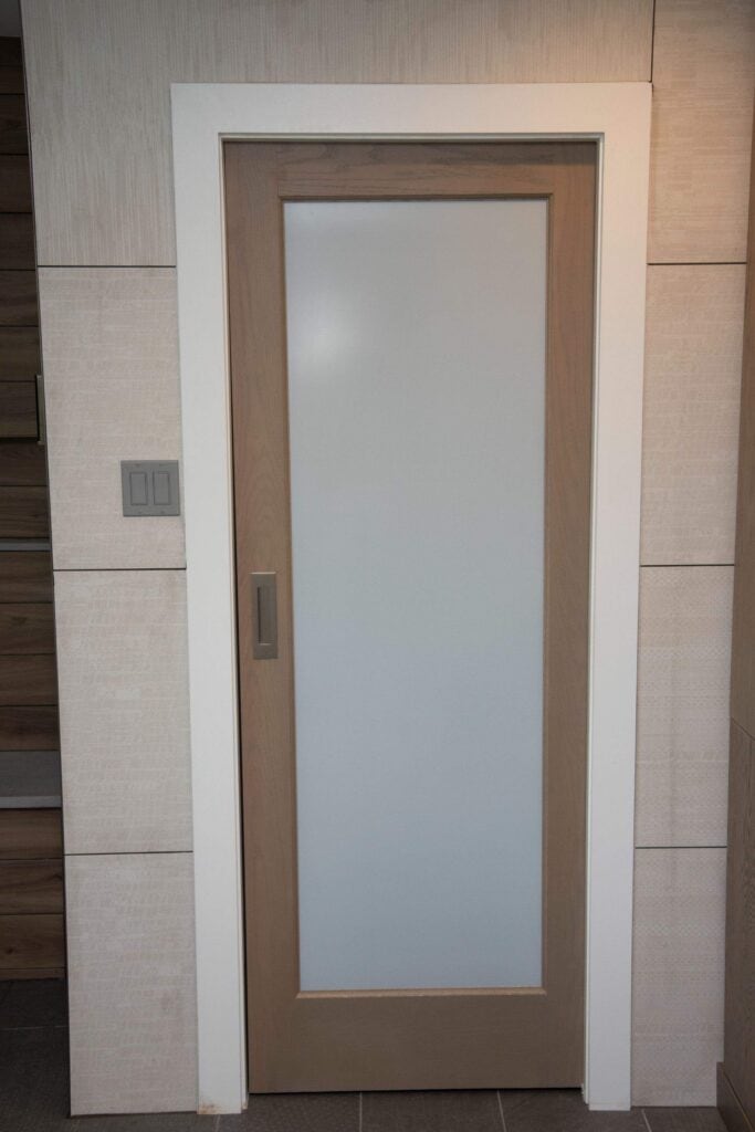 A wooden door with a frosted glass panel in the center, surrounded by a white frame, set in a modern interior wall with light-colored tiles and two light switches on the left.