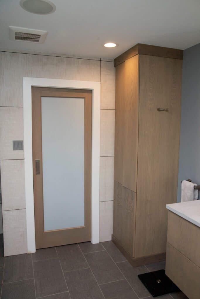 A modern bathroom with beige and gray tile flooring, light wood cabinetry, a frosted glass door with a wood frame, white-panel walls, and rolled towels on a counter.