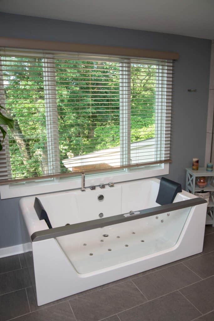 A modern white bathtub with built-in jets and black headrests sits in front of a large window with white blinds, offering a view of green trees outside. The bathroom has gray tiled floors and blue-gray walls.