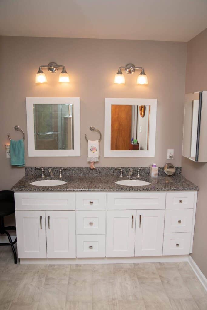 A modern bathroom with a double sink vanity, white cabinets, granite countertop, two mirrors, wall-mounted lights above each mirror, and neutral-colored walls and floor. Towels and toiletries are neatly arranged.