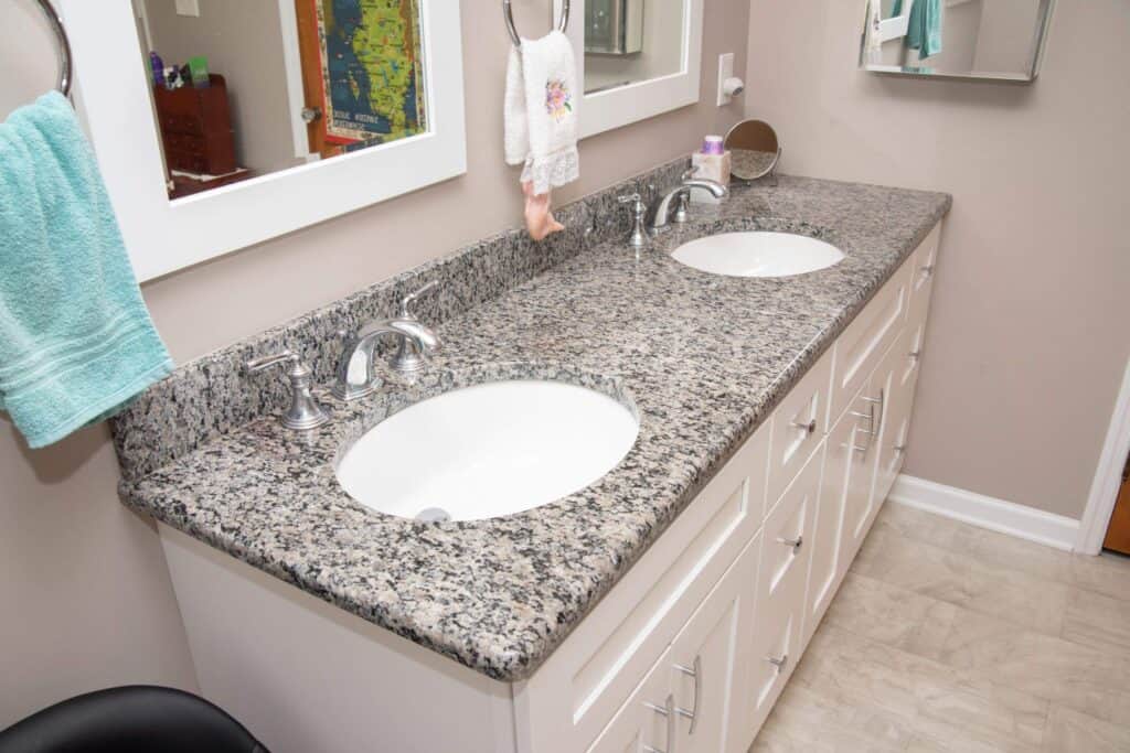 A bathroom with a granite countertop featuring two sinks, silver faucets, white cabinets below, mirrors above, and towels hanging on the wall. The floor is light-colored tile.