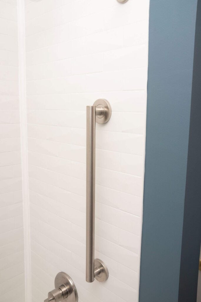 A stainless steel grab bar is mounted vertically on a white tiled shower wall, with partial view of a blue painted edge and a shower control handle at the bottom.