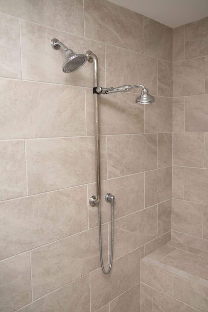 A tiled shower features two chrome showerheads attached to a vertical metal pipe, one overhead and one handheld, with a flexible hose. The beige tiles are arranged in a staggered pattern.
