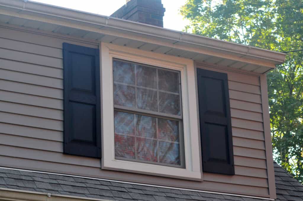 A close-up of a house dormer with beige siding, a window with white trim, black shutters on both sides, and trees reflected in the glass. Sunlight filters through the leaves in the background.