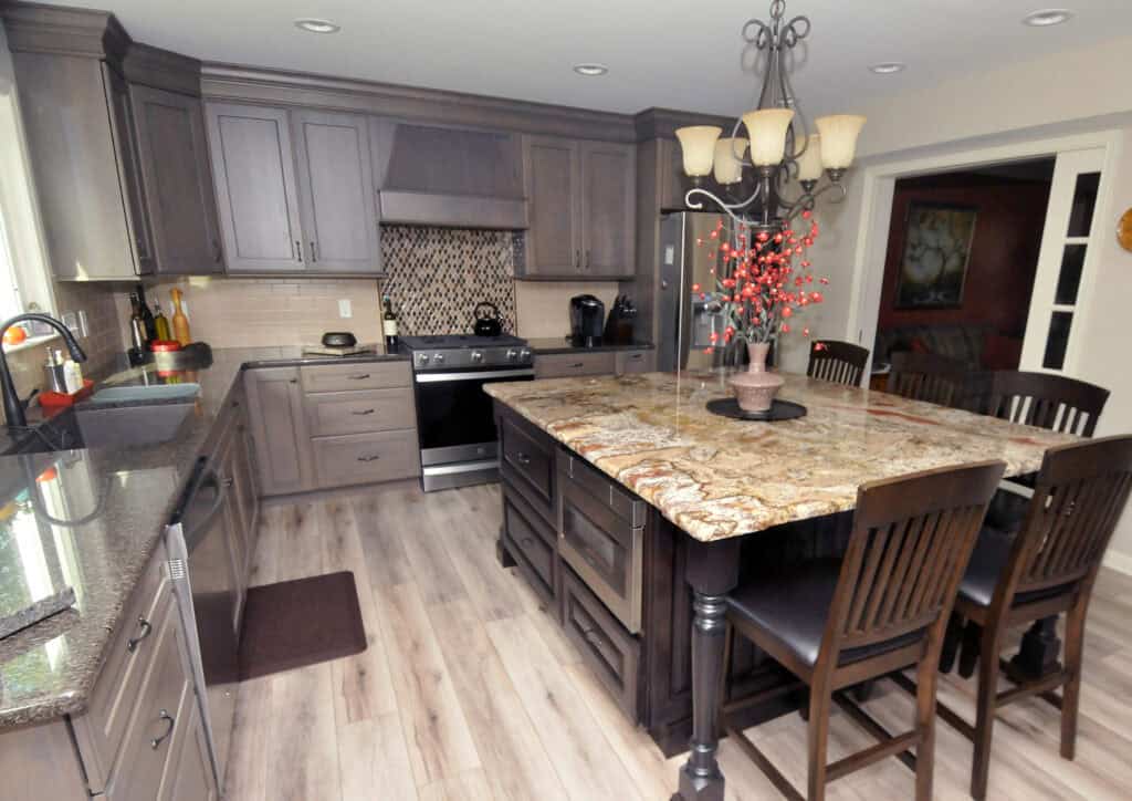 A modern kitchen featuring gray cabinets, a patterned tile backsplash, granite countertops, a large island with four chairs, and a decorative chandelier above the island.