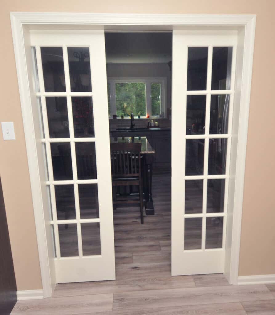 White French doors with glass panes slide open to reveal a kitchen with an island, chairs, and a window in the background. The room has light wood flooring and beige walls.