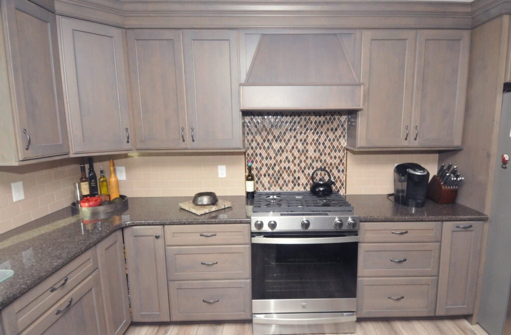 Modern kitchen with gray cabinets, a stainless steel oven and stove, granite countertops, a decorative tile backsplash, and various kitchen items including a coffee maker, knife block, and bottles on the counter.