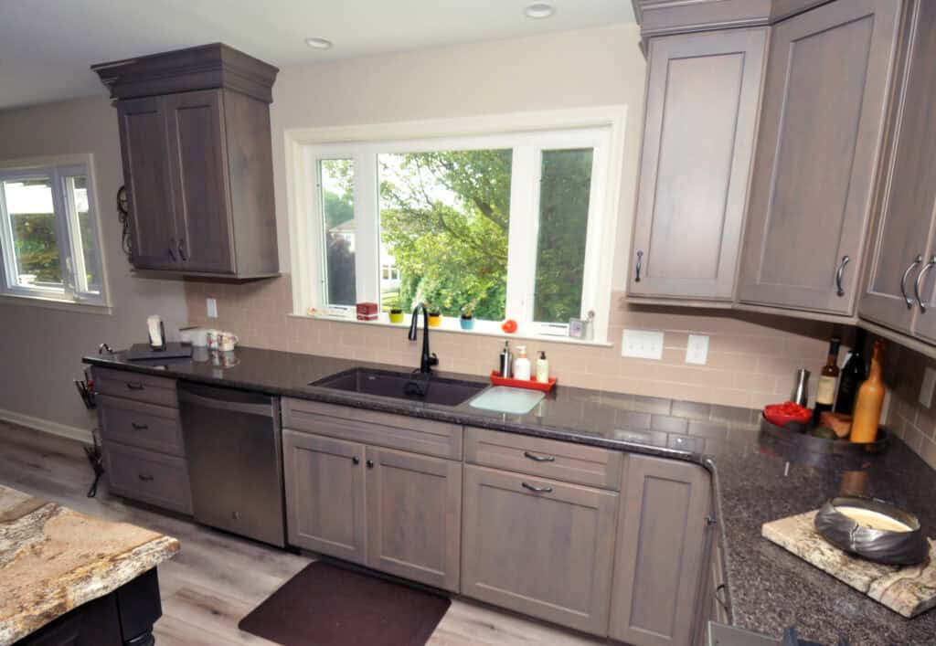 Modern kitchen with gray cabinets, black countertop, built-in dishwasher, and a large window above the sink letting in natural light. Various kitchen items and decor are arranged on the counters.