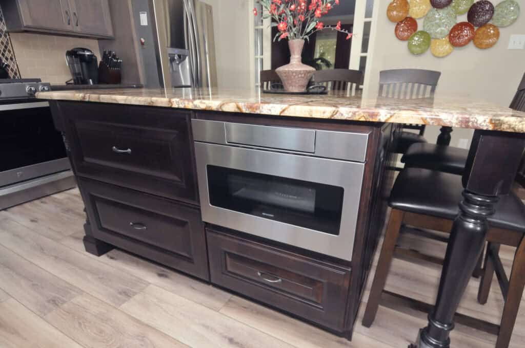 A modern kitchen island with a built-in stainless steel microwave drawer, dark cabinetry, and a marble countertop. A vase with red flowers sits on top, and chairs line one side of the island.