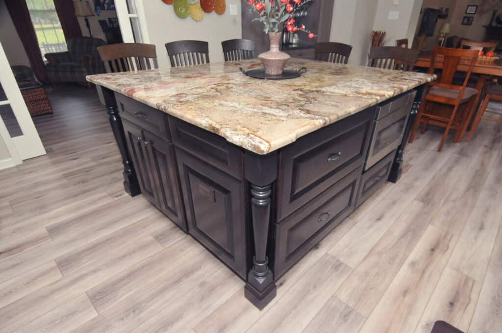 A large kitchen island with a marble countertop and dark wood cabinets, set on light wood flooring in an open dining area. A decorative centerpiece sits atop the island.
