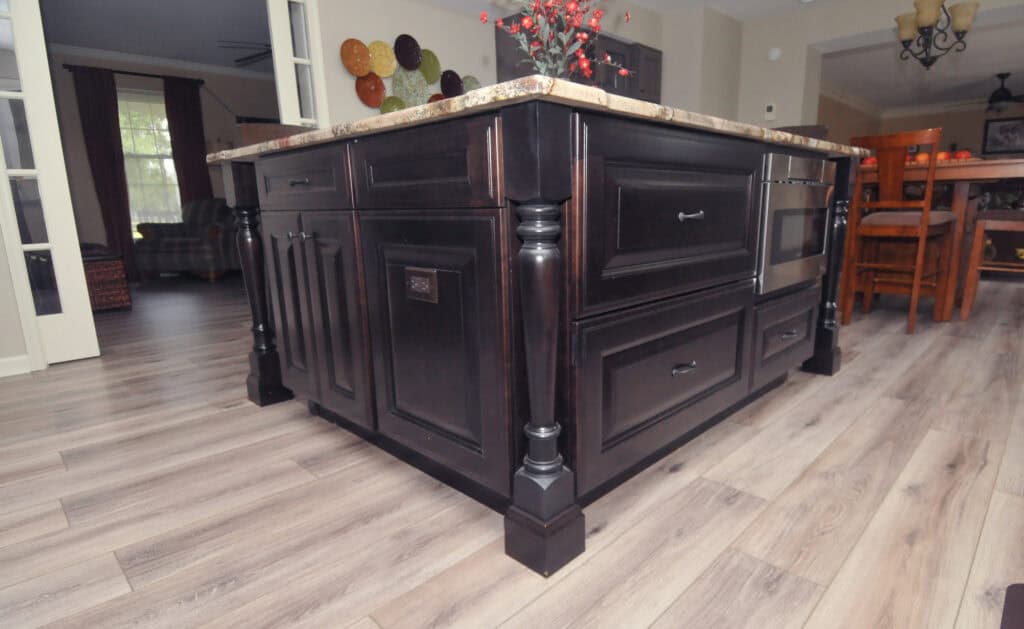 A close-up of a dark wood kitchen island with decorative legs, drawers, and a marble countertop, set on light wood flooring in a modern, open kitchen.