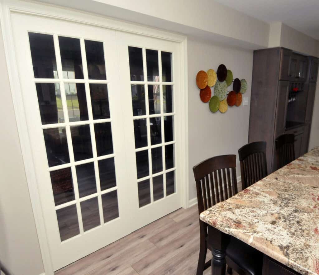A pair of white French doors with glass panes lead to another room. Next to the doors, there is wall art made of colorful circular pieces. Dark wooden chairs and a granite countertop are visible in the foreground.