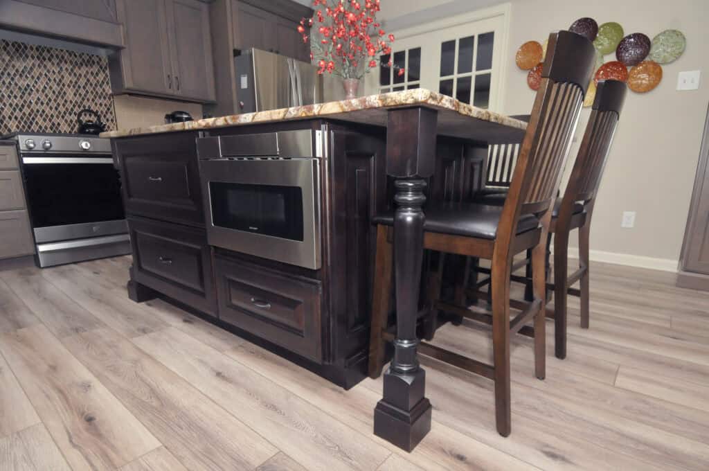 A kitchen island with a marble countertop and built-in microwave, dark wood cabinets, and two high-backed wooden chairs, set on light wood flooring with a stove and decorative wall plates in the background.