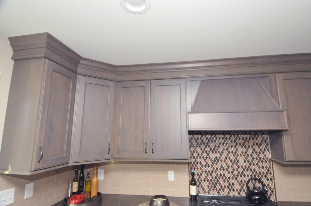 Upper kitchen cabinets with a wood finish are installed above a countertop. The backsplash features a diamond tile pattern above the stove, and various bottles and a bowl of tomatoes are on the counter.