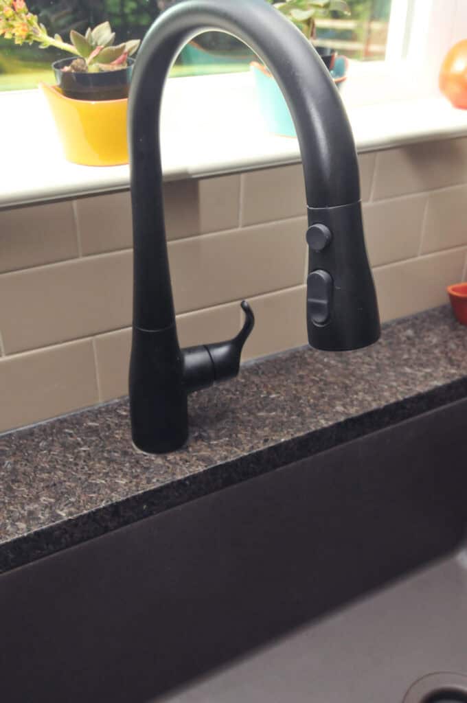 A black kitchen faucet with a curved spout and spray buttons is installed on a dark countertop next to a deep sink. Light-colored tiles and potted plants are visible on the windowsill in the background.