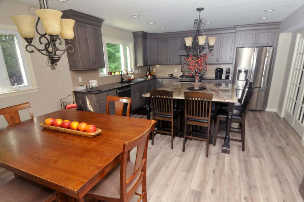 A spacious kitchen and dining area with wooden cabinets, a central island with chairs, stainless steel appliances, and a wooden dining table with a tray of apples. Two chandeliers hang from the ceiling.
