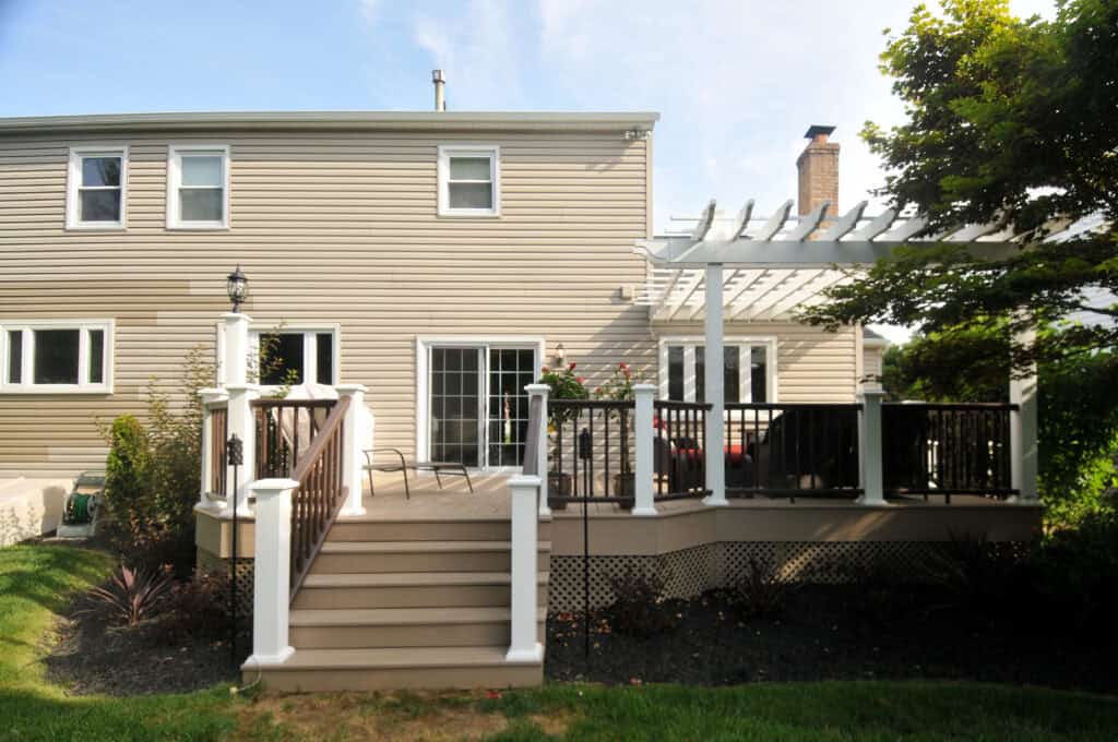 A beige two-story house with a raised wooden deck, white railings, and a pergola. The deck has outdoor chairs and is surrounded by landscaped plants and green grass.