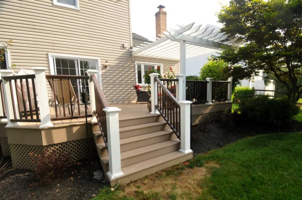 A wooden backyard deck with railings and stairs leads to a house. The deck has white posts and a pergola, with a green lawn, shrubs, and a tree nearby. Outdoor furniture is visible on the deck.