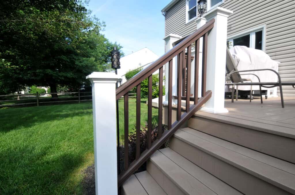Tan wooden deck steps with white and brown railing, leading up to a house with beige siding. There is green grass, trees, and patio furniture visible in the background.