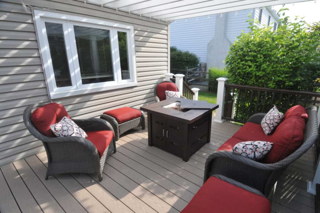 A cozy patio with wicker chairs featuring red cushions and patterned pillows surrounds a square fire pit table on a wooden deck, next to a house with gray siding and a window. Lush greenery lines the edge.