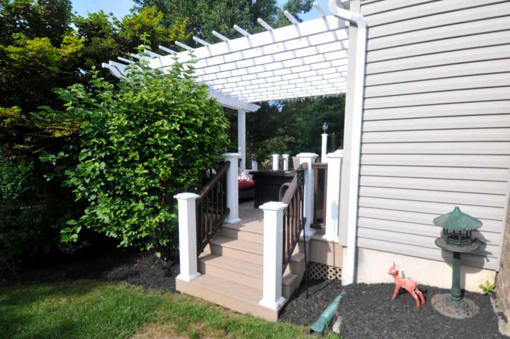 A wooden deck with stairs and white railings features a pergola overhead, attached to a house with beige siding. Green bushes surround the deck, and a decorative lantern and deer statue sit on the ground nearby.