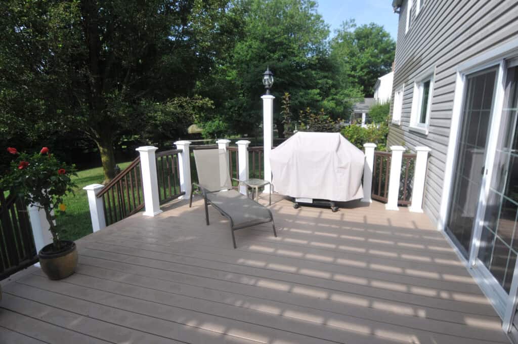 A wooden deck with white railings, a lounge chair, a covered grill, and a potted rose plant. The deck overlooks a green yard with trees and is attached to a house with sliding glass doors.