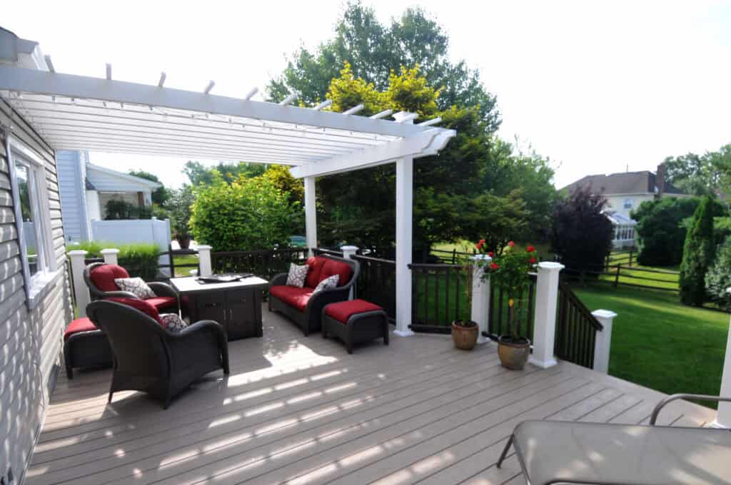 A spacious backyard deck with a white pergola, black railings, and red cushioned wicker furniture. Potted plants decorate the space, and green lawns with trees are visible in the background.