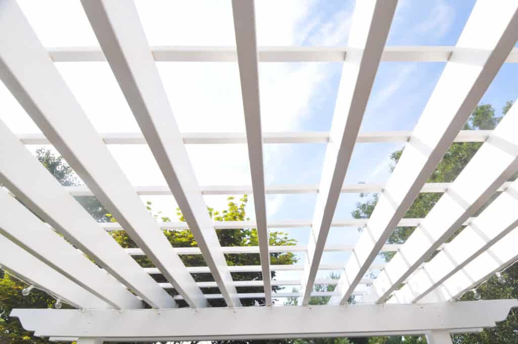 View looking up at a white pergola with evenly spaced beams, set against a blue sky with some clouds and green tree branches visible through the gaps.