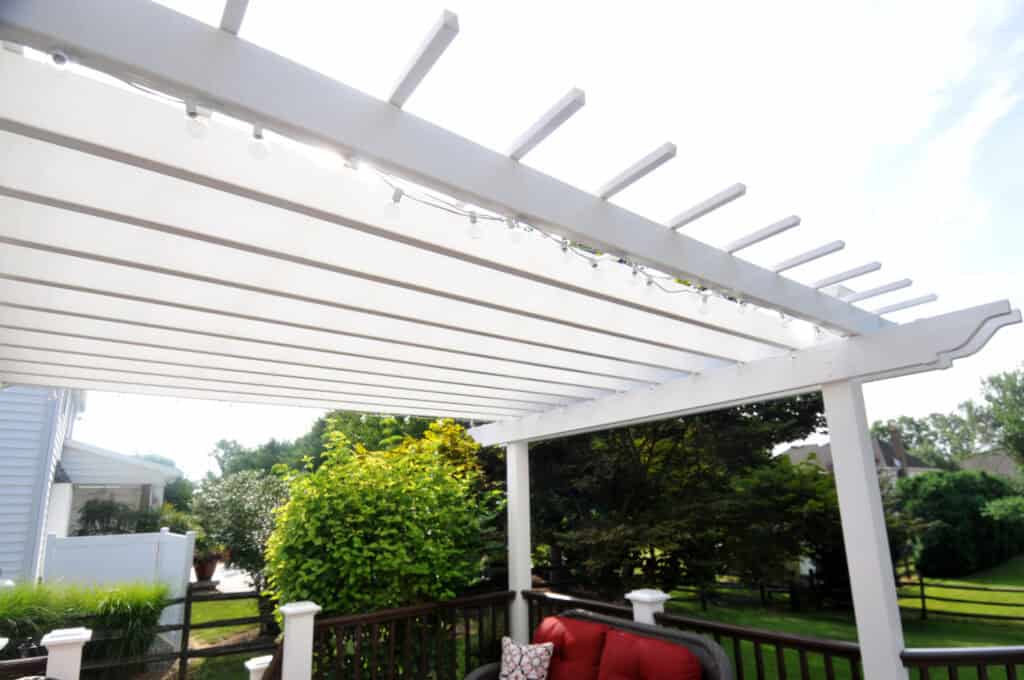 A white pergola with string lights hangs over a patio area furnished with a red cushioned chair. Green trees and bushes are visible in the background on a sunny day.