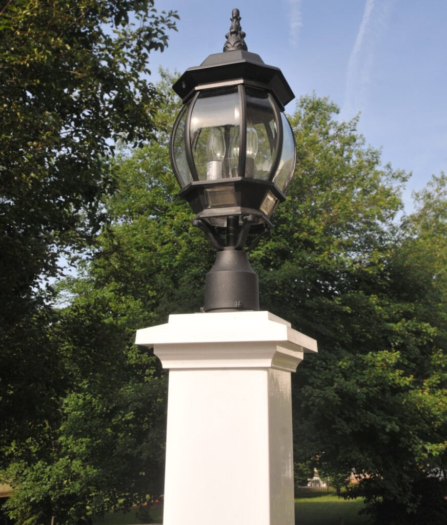 A black lantern-style outdoor light fixture mounted on a white square post, surrounded by green trees under a clear sky.