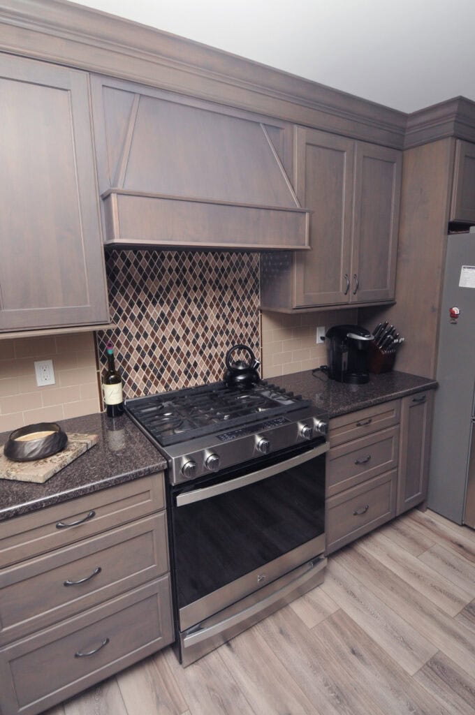 A modern kitchen with gray wood cabinets, a stainless steel gas stove, diamond-pattern tile backsplash, countertop appliances, a knife block, a bottle of wine, and light wood flooring.