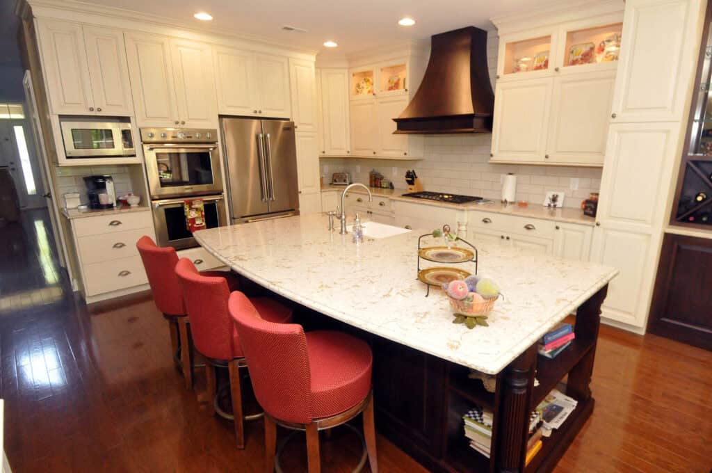 A spacious kitchen with cream cabinets, stainless steel appliances, a large marble island with a sink, three red upholstered barstools, a dark wood range hood, and hardwood floors.