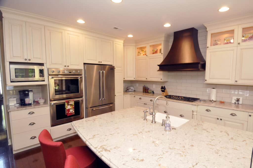 Modern kitchen with cream-colored cabinets, stainless steel appliances, a large marble island with a sink and faucet, a red chair, and a dark range hood above a gas stove. Recessed lighting brightens the space.
