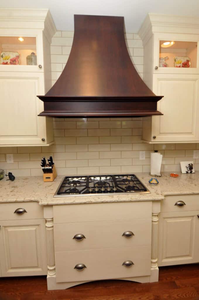 A kitchen with a beige marble countertop, a built-in gas stovetop, cream-colored cabinets, and a large dark wood range hood above white subway tile backsplash. Utensils and a knife block are on the left.