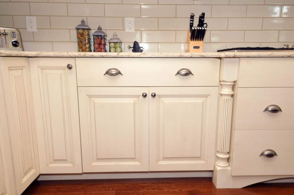 A kitchen with cream-colored cabinets and drawers, a granite countertop, jars of pasta and beans, a knife block, and a white tile backsplash. The lower cabinets have silver handles and the floor is wood.