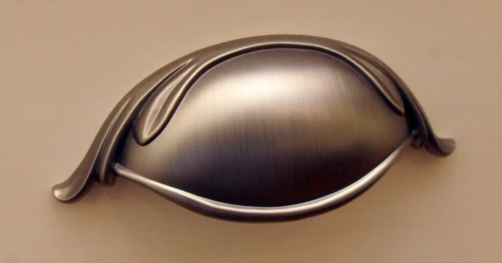 A metallic, oval-shaped cabinet drawer pull with a brushed finish, featuring an elegant, curved design on its edges, mounted on a light-colored surface.