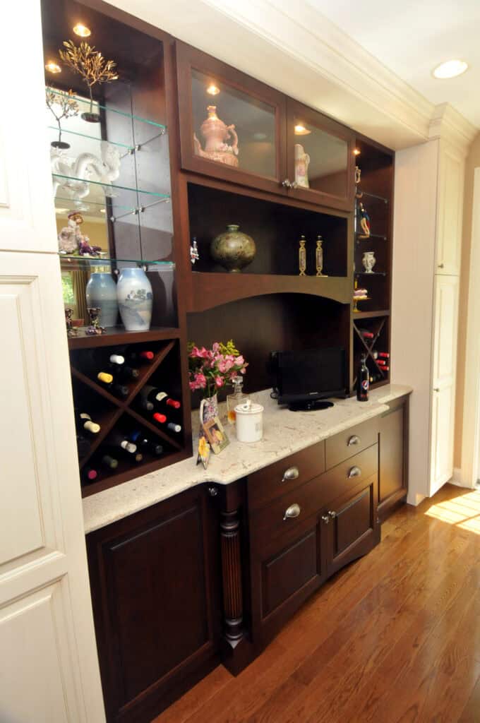 A built-in home bar with dark wood cabinets, glass shelves displaying decorative items, wine bottles stored in cubby holes, a counter with flowers, a photo frame, and a small appliance, all set on a wooden floor.