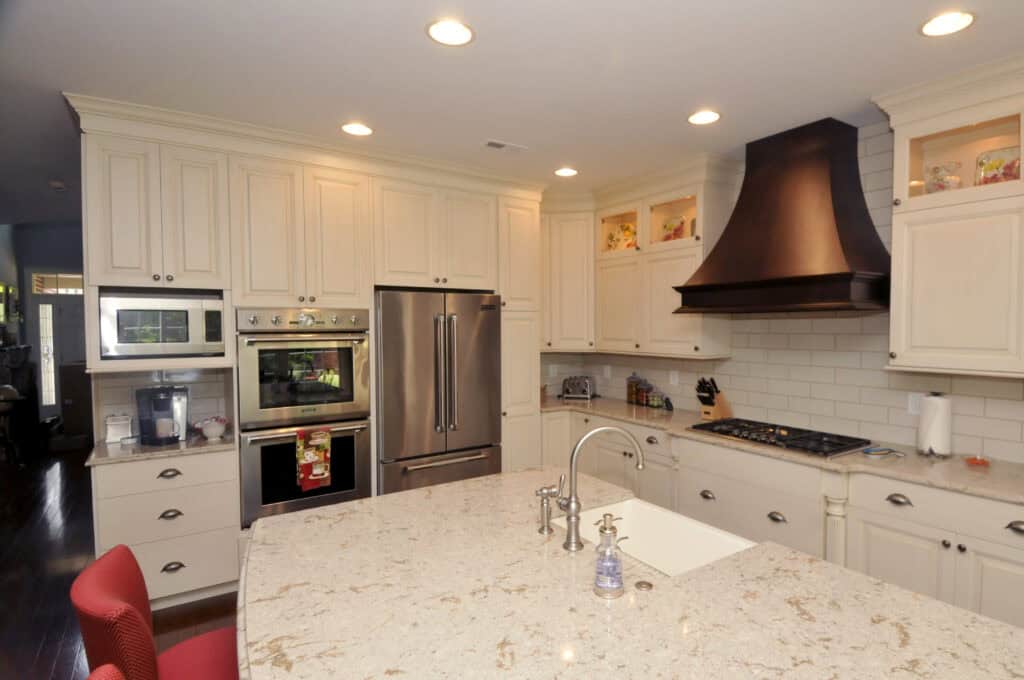 A modern kitchen with cream cabinets, stainless steel appliances, a brown vent hood, a marble island with a sink, and recessed lighting. Red chairs are partially visible at the island.