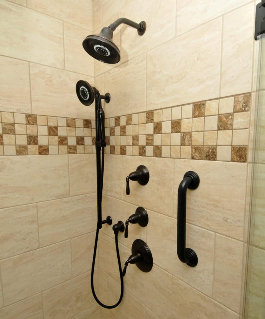 Shower with beige tiles, a dark bronze rainfall showerhead, a handheld showerhead, two control knobs, and a grab bar. Decorative square tile border runs horizontally through the middle.
