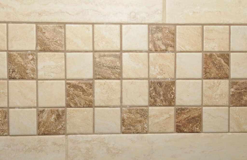 A close-up view of a tiled wall featuring small square tiles in beige and brown tones, arranged in a grid pattern, with a horizontal strip of darker tiles running through the middle.