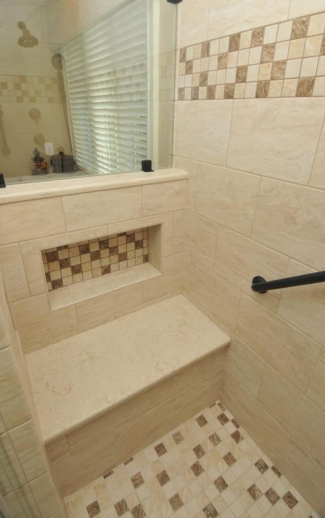 A tiled shower features a built-in bench, a recessed shelf with decorative mosaic tiles, beige wall tiles, a grab bar, and a window with blinds providing natural light.