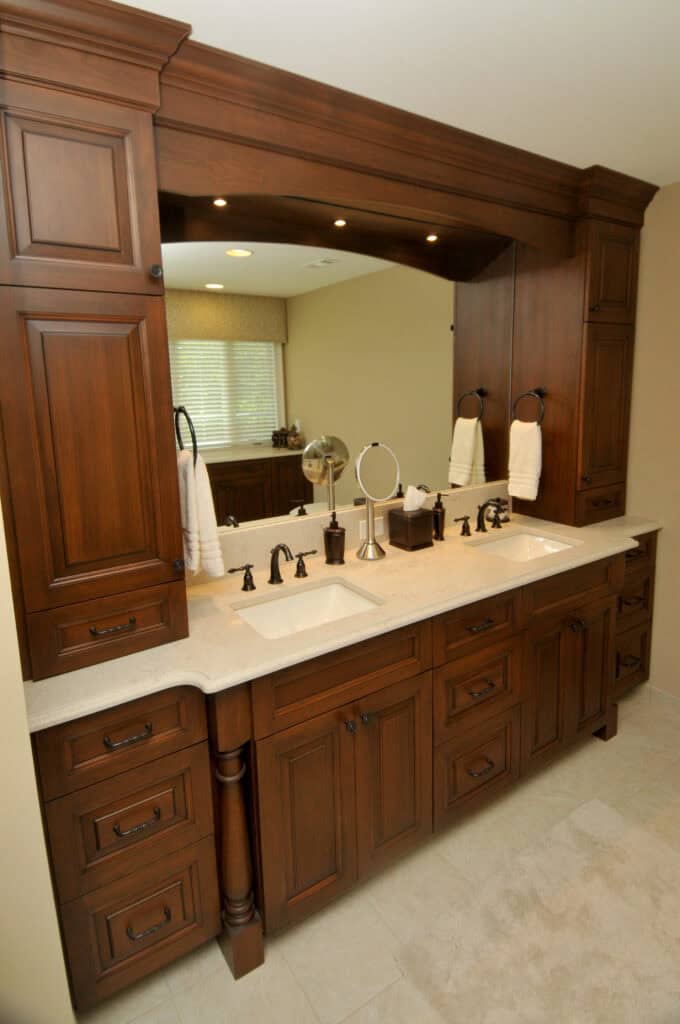 Double bathroom vanity with rich dark wood cabinets, white countertop, two sinks, bronze fixtures, large mirror, and towel holders, set against a beige wall and light-colored tile floor.