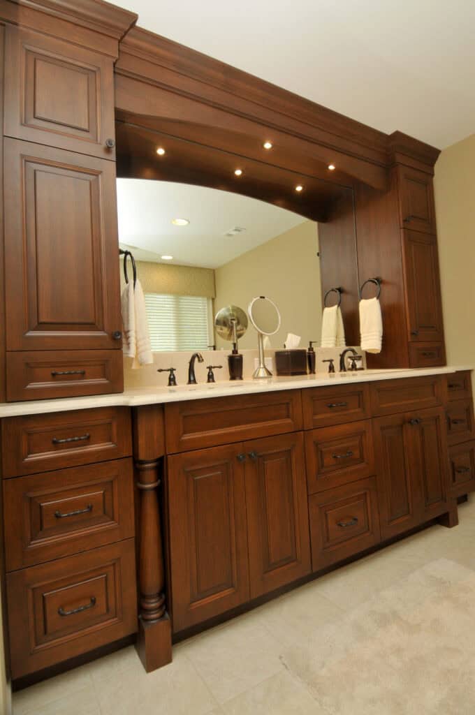 Spacious bathroom vanity with dark wood cabinets, double sinks, marble countertop, large framed mirror, two towel rings, faucets, and vanity lights above. Light tile flooring and a window with blinds in the background.